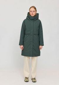 Grüne wasserdichte Parka mit hohem Kragen, Seitentaschen und tailliertem Schnitt. Getragen über hellen, weiten Hosen und gemusterten Schuhen.