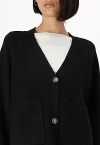 Cardigan nero realizzato in tessuto testurizzato, con scollatura a V, due tasche frontali e due bottoni con una leggera lucentezza.