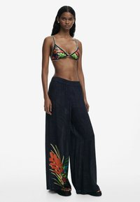 Bikini-Oberteil mit bunten Blattmustern auf schwarzem Grund, kombiniert mit weiten dunklen Hosen mit floralen Akzenten und einem elastischen Bund.