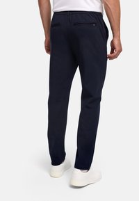 Férfi, sötétkék nadrágban és fehér tornacipőben, háttal áll sima fehér háttér előtt.