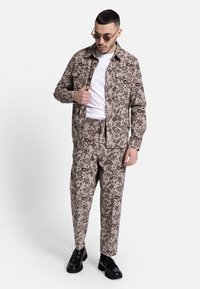 Σετ από καφέ πουκάμισο και παντελόνι με μαύρο λουλουδάτο σχέδιο, διαθέτοντας δύο τσέπες στο στήθος, φορεμένο πάνω από ένα λευκό T-shirt. Μαύρα παπούτσια ολοκληρώνουν το ντύσιμο.