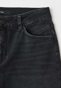 Jean en denim noir en gros plan montrant la ceinture, le bouton, la passants de ceinture, la poche avant et la petite poche à monnaie avec des détails de couture visibles.