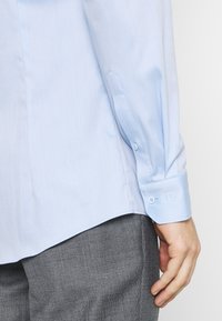 Jasnoniebieska koszula z tkaniny o fakturze, z mankietami na guziki i zaokrąglonym dołem, połączona z szarymi spodniami o fakturze.