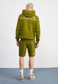 Felpa verde oliva con logo sulla schiena, abbinata a pantaloni corti coordinati. Caratteristiche includono una tasca a marsupio e tessuto elasticizzato. Calzature casual.