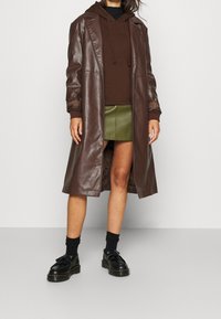 Long brown leather coat over a dark brown hoodie, paired with a green skirt and black shoes. Features include both smooth and textured materials.