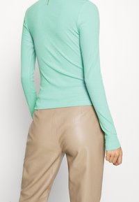 Ljusgrön långärmad topp med ribbad struktur, kroppsnära design och dragkedja i ryggen, tillsammans med beiga blanka byxor.