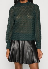 Transparent, mörkgrön spetsblus med ett texturerat diamantmönster och puffärmar, kombinerad med en svart plisserad läderskjol.