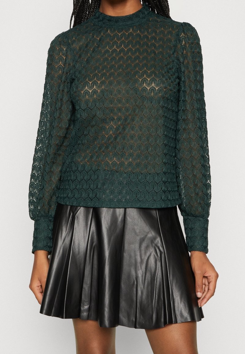 Transparent, mörkgrön spetsblus med ett texturerat diamantmönster och puffärmar, kombinerad med en svart plisserad läderskjol.