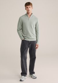 Svijetlozeleni džemper sa zatvaračem i teksturiranom površinom, upareni s crnim trapericama i sivim tenisicama, stojeći na neutralnoj pozadini.