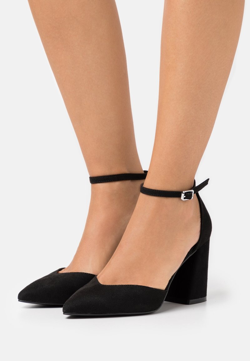 Jambes portant des escarpins à talons blocs en daim noir à bouts pointus, avec des brides autour de la cheville et des boucles argentées, sur un fond blanc.