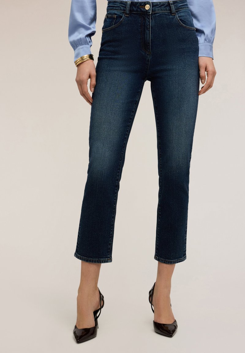 Vrouw die donkerblauwe cropped jeans draagt, een lichtblauwe blouse, zwart puntige hakken en gouden armbanden aan één pols.
