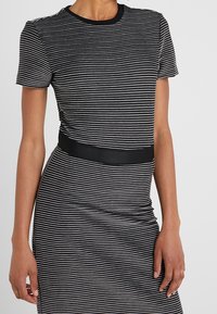 Femme portant une robe ajustée à rayures horizontales noires et blanches avec manches courtes et une ceinture noire unie à la taille, sur un fond uni.