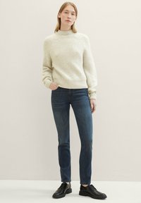 Suéter crema de canalé con cuello alto, combinado con jeans ajustados de color azul oscuro y zapatos negros de cordones, de pie frente a un fondo neutral.