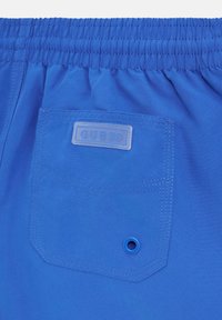 Pantaloni corti blu con vita elasticizzata, una tasca sul retro con un logo in plastica "GUESS" e un piccolo dettaglio in occhio di bue. Tessuto dalla consistenza liscia.