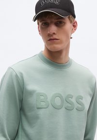 Világoszöld pulóver texturált "BOSS" logóval, kerek nyakkivágással és látható varrással. Fekete sapka hálós betétekkel és ívelt karimával.