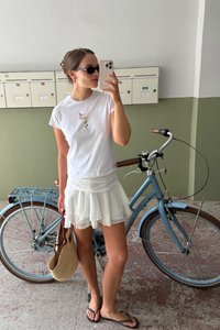 Baltas T-krekls ar kokteiļu grafiku, kārtots balts svārks, čības un austs soma. Zils velosipēds ar brūniem akcentiem blakus.