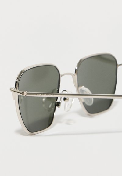 Γυαλιά ηλίου σε σχήμα τετράγωνου με ασημένια μεταλλικά πλαίσια και σκουρόχρωμους φακούς, με ρυθμιζόμενα μαξιλαράκια στη μύτη και το όνομα "McQueen" χαραγμένο στο πλαϊνό μέρος.