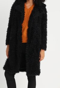 Sort pelsjakke i imiteret pels med lange ærmer og åben front, der har en tekstureret, shaggy overflade og en afslappet pasform.