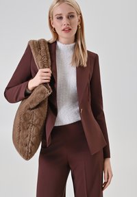 Brauner maßgeschneiderter Blazer und Hose, kombiniert mit einem weißen strukturierten Pullover und einer großen, braunen Pelztasche mit geflochtenem Muster.