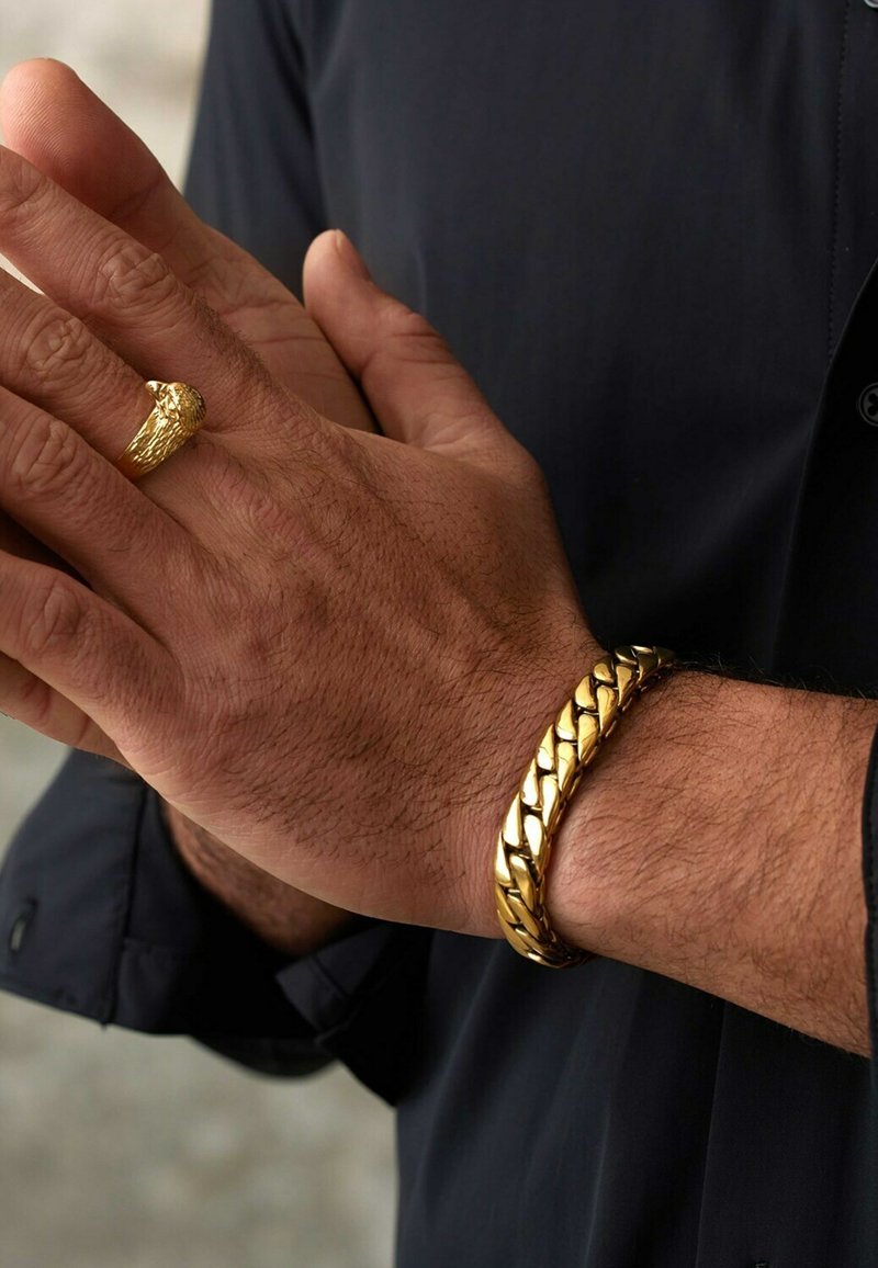 Gouden kettingarmband met in elkaar geweven schakels om de pols, vergezeld van een goudkleurige ring met een gestructureerd ontwerp in de vorm van een adelaarskop op een andere hand.