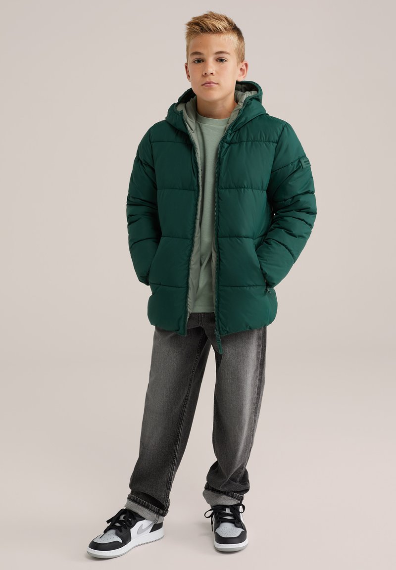 Zelena napihnjena jakna s kapuco in prešito zasnovo, nosi se preko svetlo zelene srajce. Uklaplja se s temno sivo običnimi hlačami in črnimi supergami.