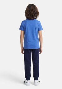 Παιδί με σγουρά μαλλιά στέκεται γυρισμένο ανάποδα, φοράει μπλε κοντομάνικο πουκάμισο, σκούρο μπλε παντελόνι και λευκά αθλητικά παπούτσια μπροστά σε ένα απλό φόντο.