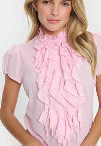 Rosa blus med en rynkad frontdesign, korta ärmar och en hög nacke med en samlad detalj. Lätt material med en slät textur.