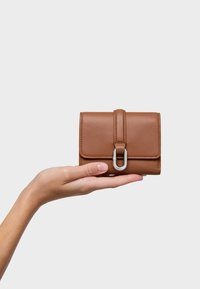 Cartera de cuero marrón con dise�ño de solapa, que presenta un adorno metálico plateado y bordes cosidos, sostenida en una mano sobre un fondo simple.