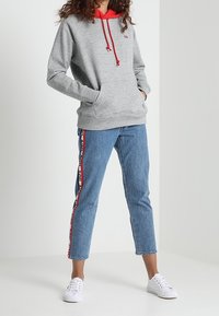 Sudadera gris con forro interno rojo y cordones, combinada con jeans azul claro que cuentan con franjas de logo rojas y negras a los lados, y zapatillas blancas.