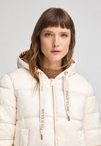 Mujer con cabello castaño claro y ojos azules que lleva una chaqueta de plumas color crema con cordones que dicen "POLO CLUB" y un suéter marrón debajo.