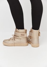 Sneakers beige alte con design lacciato, cinturino in velcro e suola in gomma testurizzata, realizzate in un materiale sintetico liscio.