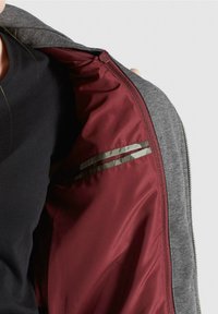 Šedá bunda s vínově červeným vnitřkem, vybavená horizontálním kapsou s reflexními páskami. Tkanina působí hladce a měkce.