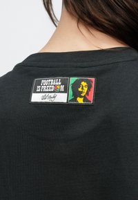 Άτομο που φοράει μαύρο πουκάμισο με ένα σήμα που γράφει "ΤΟ ΠΟΔΟΣΦΑΙΡΟ ΕΙΝΑΙ ΕΛΕΥΘΕΡΙΑ" και πολύχρωμο στυλιζαρισμένο πορτρέτο ενός ατόμου με ντραντλοκς.