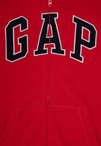 Roter Reißverschluss-Hoodie mit großen marineblauen Buchstaben, die "GAP" buchstabieren. Weicher Stoff, Kängurutaschen und gerippte Bündchen und Saum.