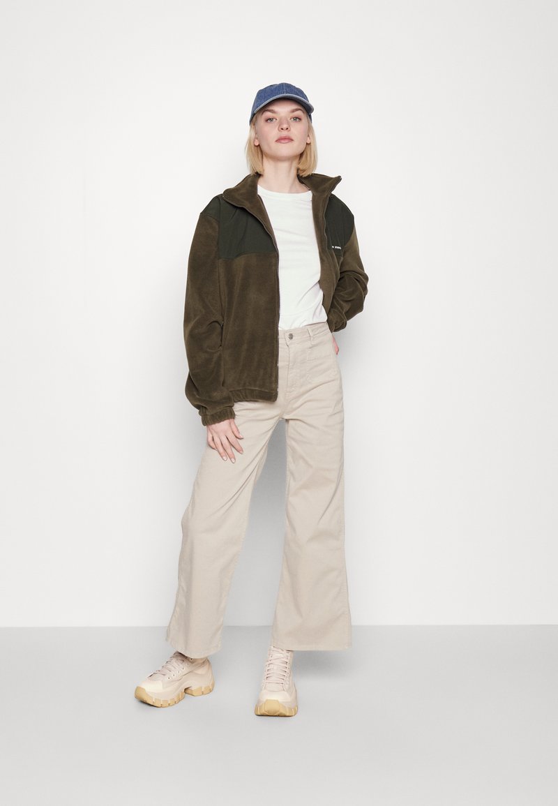 Giacca in pile di colore verde oliva e nero, abbinata a una t-shirt bianca e pantaloni beige a gamba larga. Sneakers chiare completano l'outfit.