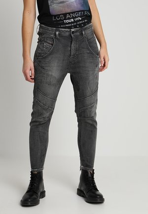 Persona con vaqueros negros ajustados y desgastados con detalles cosidos, combinados con botines negros con cordones y una camiseta negra con diseño gráfico.