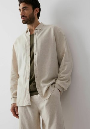 Mężczyzna z ciemnymi włosami i brodą, ubrany w rozpiętą beżową koszulę na oliwkowozielonym t-shircie i pasujących beżowych spodniach, stojący przy białej ścianie.