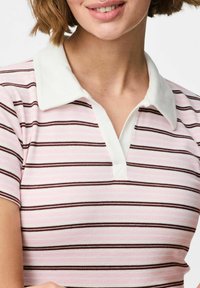 Bļodi rozā polo krekls ar plānām brūnām svītrām, ar baltu apkakli un īsām piedurknēm, izgatavots no teksturētas adītas auduma.