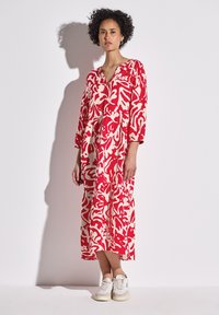 Rotes und cremefarbenes gemustertes Kleid mit V-Ausschnitt und Dreiviertelärmeln, das eine fließende Silhouette hat. Getragen mit weißen Turnschuhen.