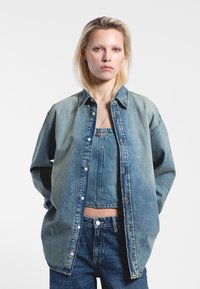 Giacca di jeans di colore azzurro chiaro, con abbottonatura frontale, colletto e vestibilità rilassata. Indossata sopra un top corto abbinato e jeans.