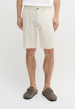 Homme portant un short en velours côtelé beige, une chemise blanche et des chaussures grises sans lacets, debout devant un fond clair uni.