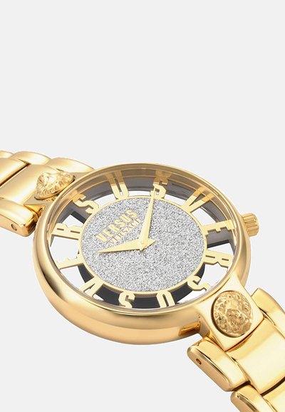 Gold-toned watch with a textured silver dial, bold cut-out letters, and lion head accents on the bracelet. Circular case design.