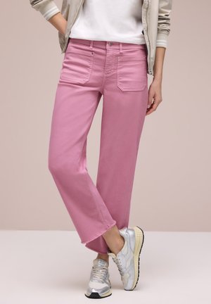 Personne portant un pantalon large rose à ourlets effilochés, des baskets blanches, un haut blanc et une veste beige clair sur un fond uni.