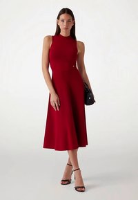 Robe rouge sans manches, longueur genou, avec un corsage ajusté et une jupe évasée. Texture lisse, détails minimaux, complétée par des accessoires noirs.