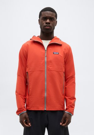 Homme portant une veste zippée orange avec capuche et poches latérales, debout devant un fond clair uni.