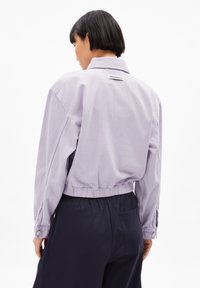Chaqueta corta de color púrpura claro con cuello, cinturilla elástica y puños abotonados. Presenta una etiqueta en la parte posterior. Tejido de algodón suave.