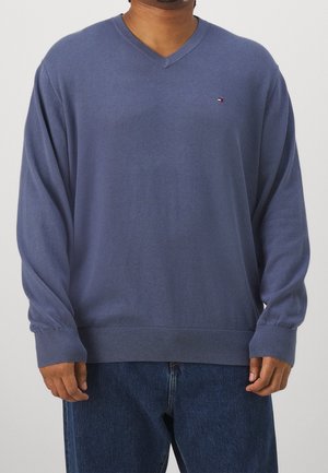 Man draagt een blauw V-hals trui met lange mouwen en donkerblauwe spijkerbroek, staand tegen een effen lichte achtergrond.