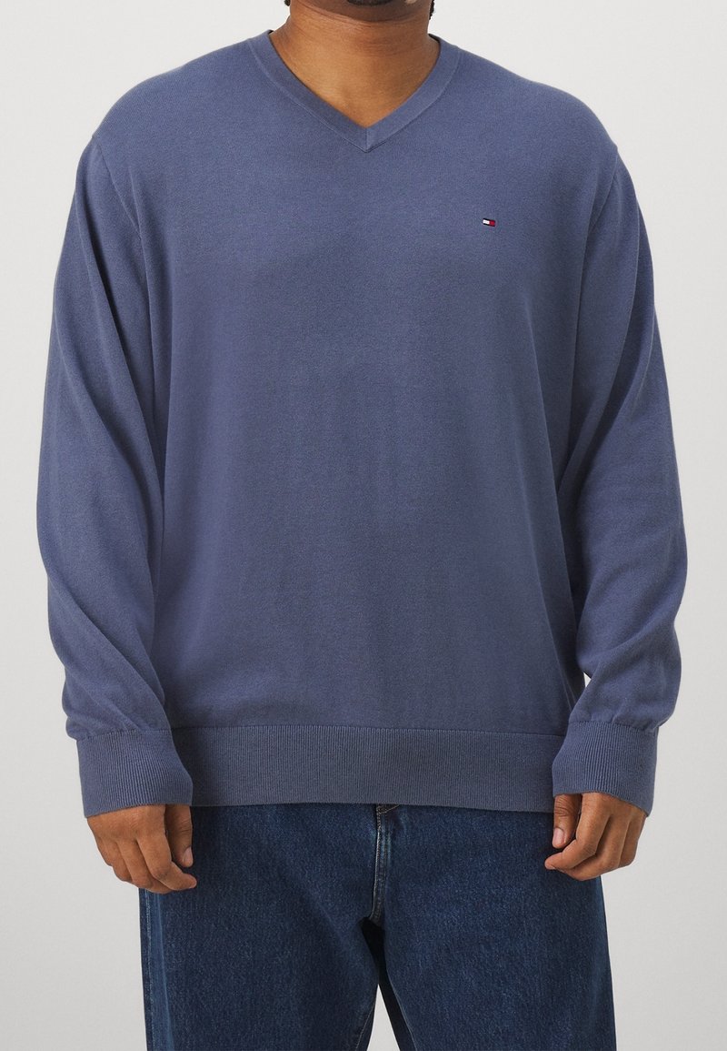 Homme portant un pull bleu à col en V à manches longues et un jean bleu foncé, debout devant un fond clair uni.