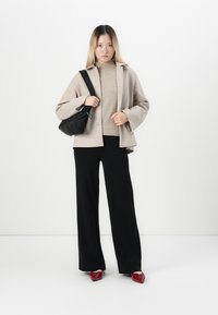 Woman standing wearing a beige jacket, light sweater, black wide-leg pants, red shoes, and carrying a black shoulder bag against a white background.