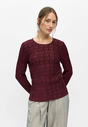 Femme aux cheveux châtain clair portant un haut bordeaux texturé à manches longues et un pantalon large gris clair, posant devant un fond blanc.